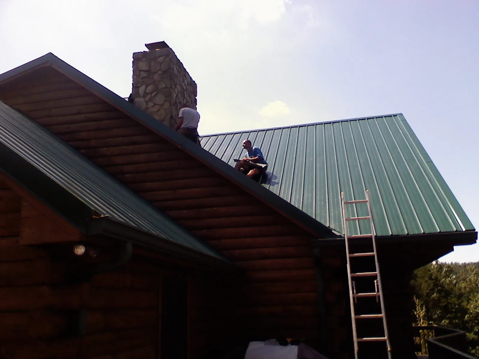 Roofers installing green metal panels beside a stone chimney on a log home in Moulton